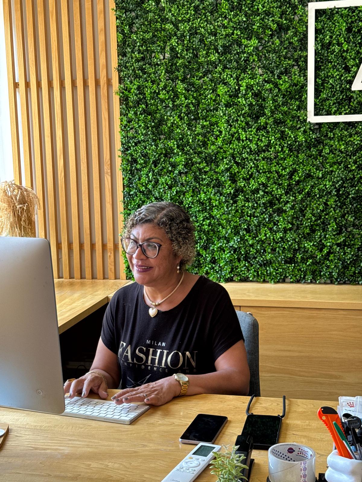 Rosângela Figueiredo sentada ao computador, em atendimento online, com parede verde ao fundo
