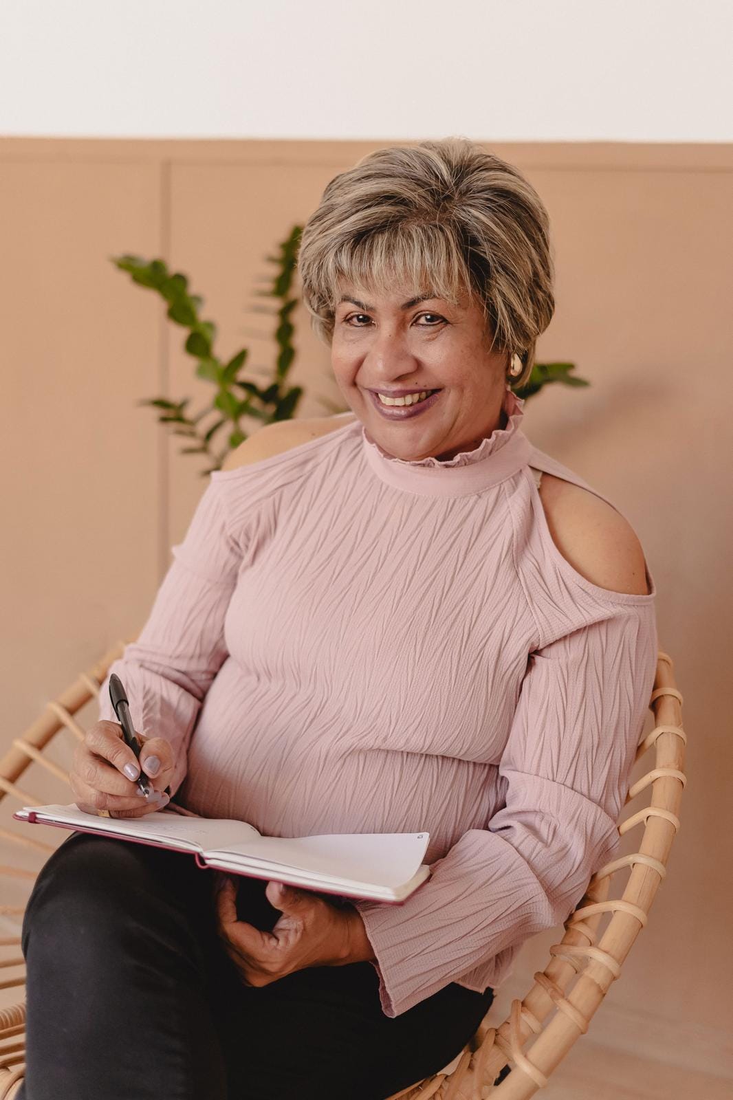 Rosângela Figueiredo, terapeuta e psicanalista, de blusa rosa, sorrindo sentada numa cadeira de vime com um caderno de anotações no colo
