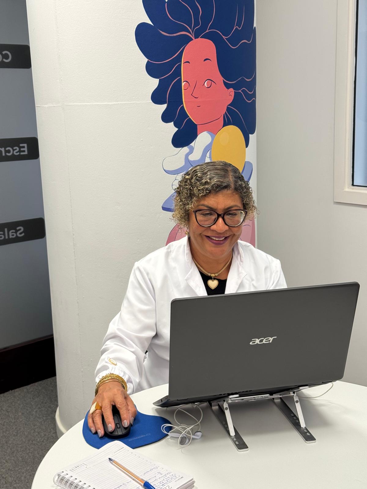 Rosângela Figueiredo em atendimento de terapia online, ao computador, com parede decorada ao fundo